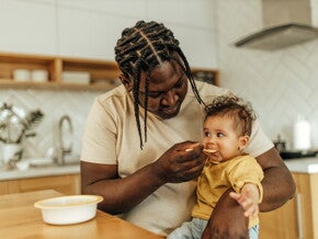Dad feeding his baby girl, sitting on his lap.