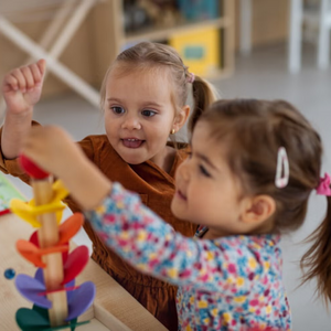 Photo of two preschool girls smiling while stacking a colorful wooden toy in a classroom
