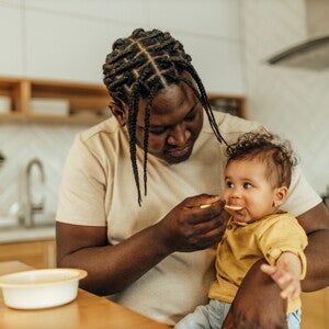 Dad feeding his baby girl, sitting on his lap.