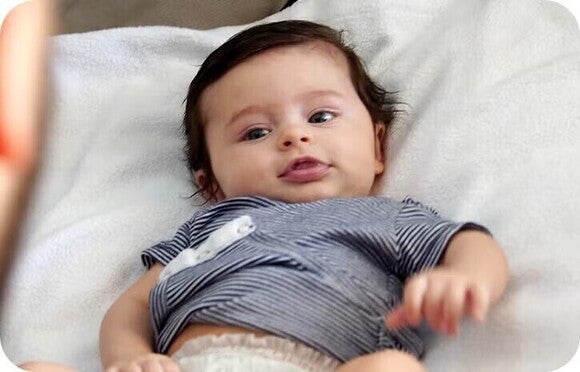 Foto van liggende baby in grijs gestreept shirt op witte deken, speels gezicht